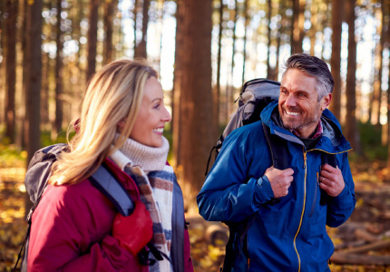 Natural Pharmaceuticals Movisan™ Collagen II Para turystów w średnim wieku w odzieży outdoorowej z plecakami, uśmiechająca się podczas wędrówki przez jesienny las.