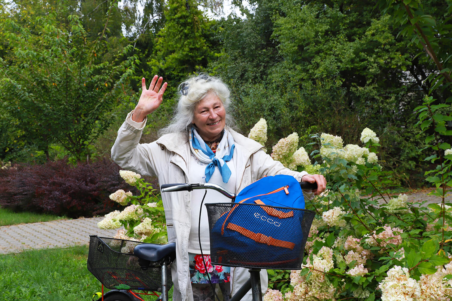 Natural Pharmaceuticals Movisan™ Collagen II Starsza kobieta uśmiechająca się i machająca stojąc obok roweru z niebieskim plecakiem w koszyku na tle zielonego ogrodu z kwitnącymi krzewami hortensji.