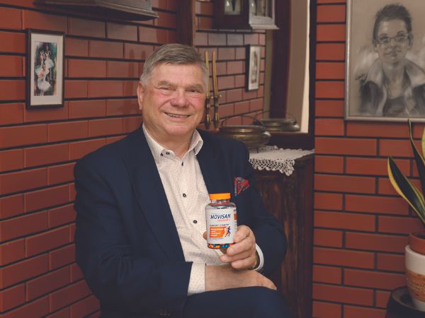 Natural Pharmaceuticals Movisan™ Collagen II Starszy mężczyzna w garniturze siedzi w pokoju z czerwonymi cegłami na ścianach, uśmiechając się i trzymając butelkę suplementu diety Movisan Collagen II.