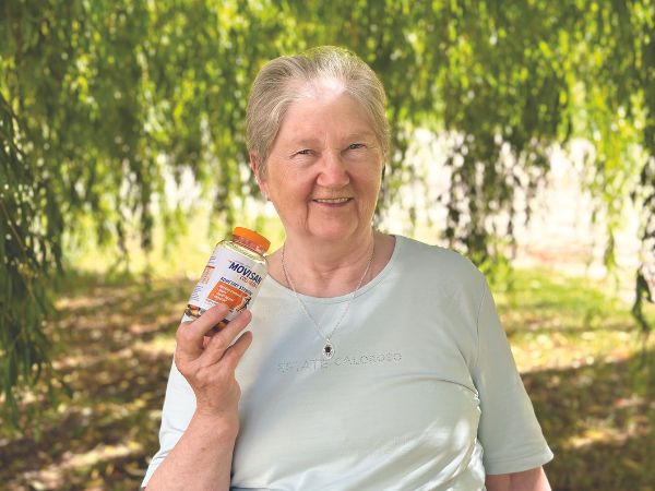 Natural Pharmaceuticals Movisan™ Collagen II Starsza kobieta uśmiechająca się i trzymająca butelkę suplementu diety Movisan Collagen II na tle zielonych drzew, symbolizująca zdrowie i wsparcie stawów.