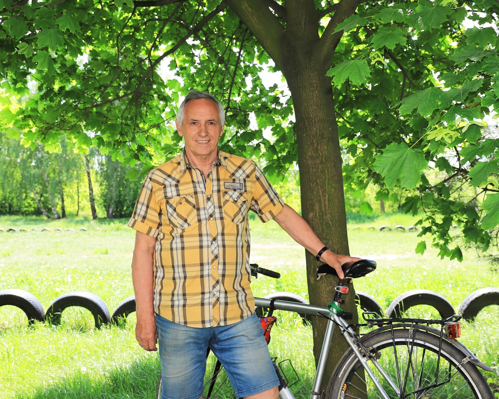 Natural Pharmaceuticals Movisan™ Collagen II Mężczyzna w żółtej koszuli w kratę i niebieskich szortach stoi przy rowerze pod drzewem w słonecznym parku z zieloną trawą i oponami w tle.