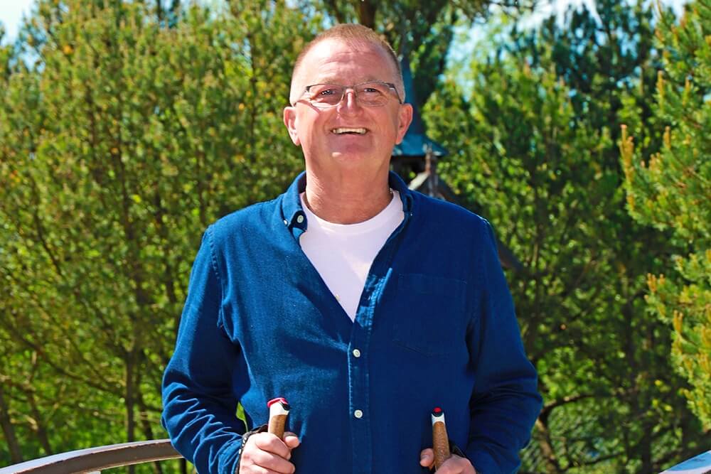 Natural Pharmaceuticals Movisan™ Collagen II Uśmiechnięty starszy mężczyzna w okularach i niebieskiej koszuli uprawiający nordic walking w zielonym, słonecznym parku.