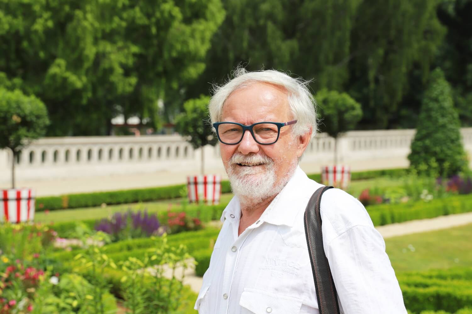 Natural Pharmaceuticals Movisan™ Collagen II Starszy mężczyzna w białej koszuli i okularach, uśmiechający się na tle zadbanego, zielonego ogrodu z kwiatami i krzewami.