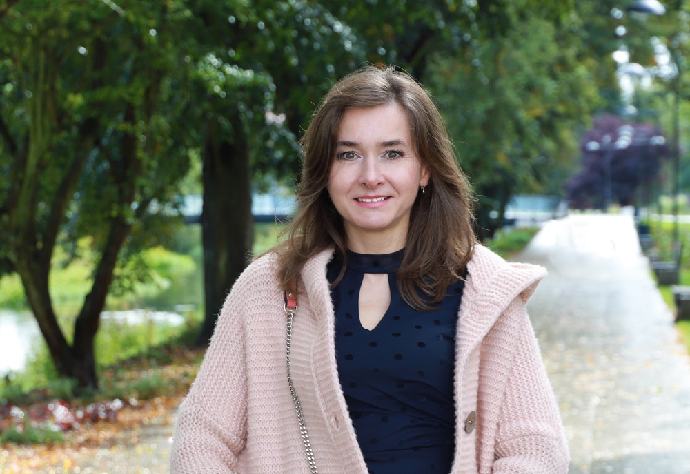 Natural Pharmaceuticals Movisan™ Collagen II Kobieta w różowym płaszczu i granatowej sukience stoi uśmiechnięta na ścieżce parkowej w otoczeniu jesiennych drzew.