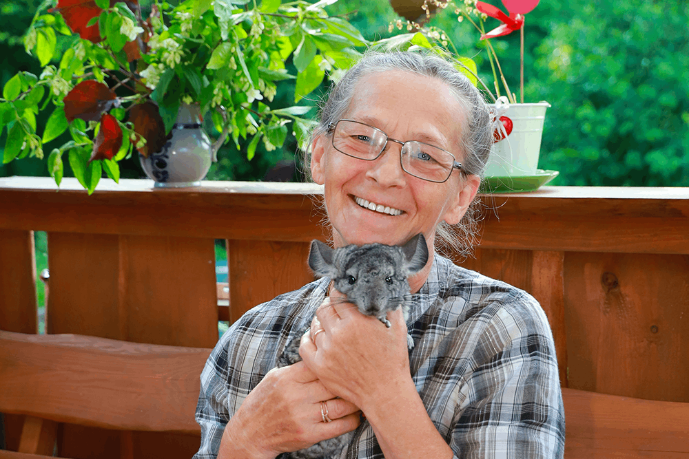 Natural Pharmaceuticals Movisan™ Collagen II Uśmiechnięta starsza kobieta w okularach trzyma w rękach szarą szynszylę na tle drewnianego ogrodzenia i zielonych roślin.