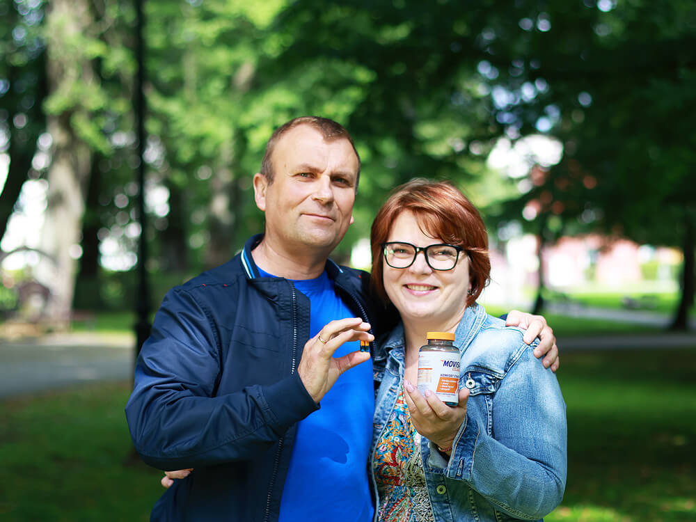 Natural Pharmaceuticals Movisan® Collagen II Para stoi w parku i uśmiechając się, trzyma suplement diety Movisan® Collagen II wspierający zdrowie stawów.